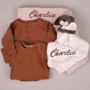 Personalized baby gift set with brown ribbed rompers, beige knitted blanket, and puppy comforter embroidered “Charlie.”