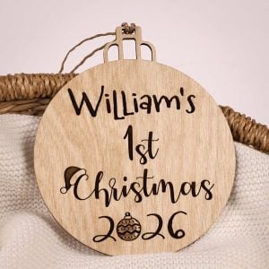 Round birch wood personalised 1st Christmas bauble disc with laser engraved name, year and Santa hat Christmas ornament design on wicker basket.