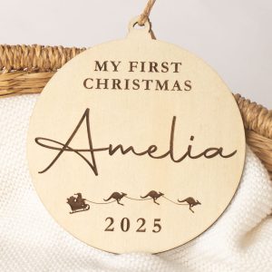 Wooden baby disc reads "My First Christmas Amelia 2025" with kangaroo sleigh engraving design.