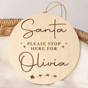 **Alt Text (15 words):** Wooden baby disc reads "Santa Please Stop Here For Olivia" with kangaroo sleigh engraving below.