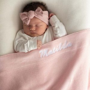 A baby laying under a large pink knitted blanket personalised with the name Matilda.