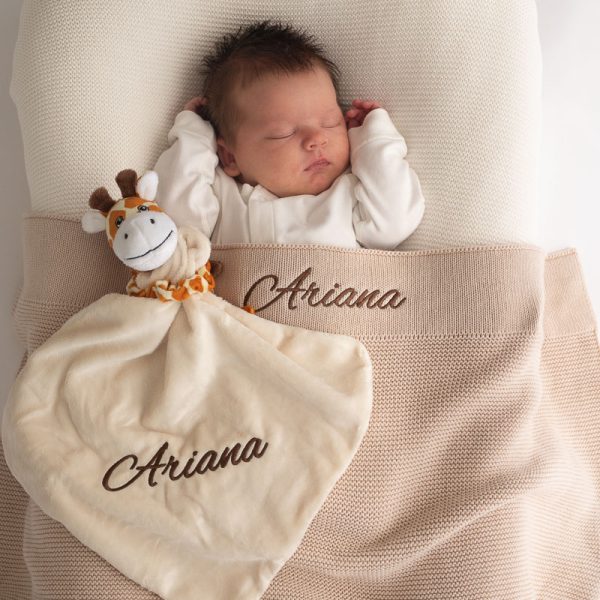 A baby laying under a beige knitted baby blanket & giraffe comforter both embroidered with the name Arianna.