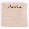 Folded beige personalised knitted baby blanket with brown embroidered script name on white background.