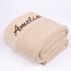 Beige diamond knitted personalised baby blanket rolled and folded with brown embroidered script name on white background.