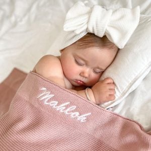 Personalised blush pink knitted blanket with Mahloh embroidered in white on sleeping baby wearing white bow headband