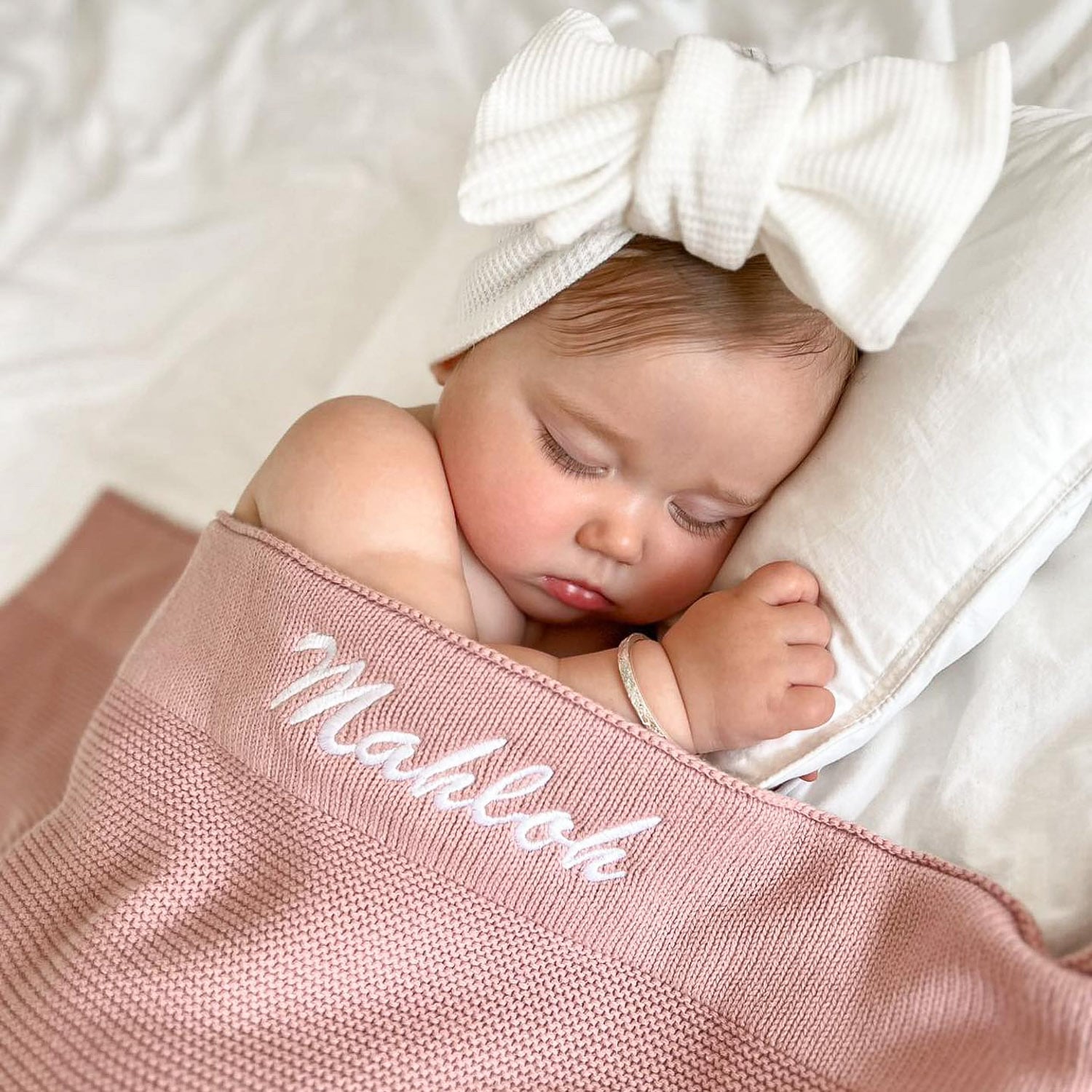 Personalised blush pink knitted blanket with Mahloh embroidered in white on sleeping baby wearing white bow headband