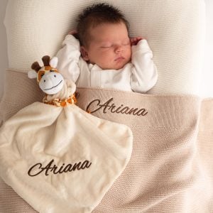 A baby laying under a beige knitted baby blanket & giraffe comforter both embroidered with the name Arianna.