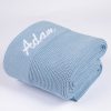 Blue knitted personalised baby blanket rolled and folded with white embroidered script name on white background.
