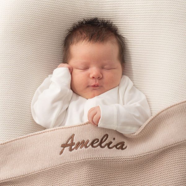 A newborn baby laying under a beige knitted blanket personalised with Amelia.