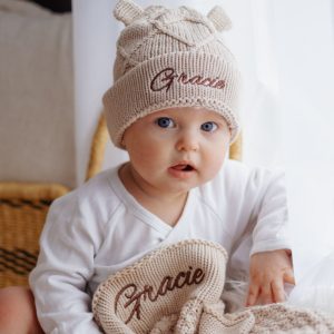 Baby wearing beige knitted hat & diamond baby blanket with name ‘Gracie’.