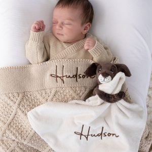 Sleeping newborn with personalised beige diamond knitted baby blanket and cream puppy comforter, both with brown embroidered script name on white bedding.