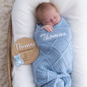 Personalised light blue diamond knit blanket with Thomas embroidered in white on swaddled newborn beside wooden nursery plaque with Thomas Tyler Grace and blue bear detail.