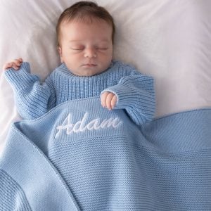 Sleeping newborn wearing a blue knitted romper wrapped in a personalised blue knitted baby blanket with white embroidered script name on white bedding.