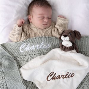 Sleeping newborn with personalised sage green diamond knitted baby blanket and cream puppy comforter, both with white and brown embroidered script name on white bedding.