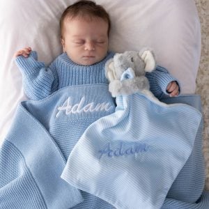 Sleeping newborn with personalised blue knitted baby blanket and blue striped elephant comforter, both with blue embroidered script name on white bedding.