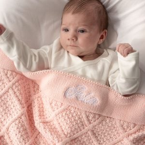 Baby lying on white bedding, wrapped in a light pink personalised knitted blanket with Isla embroidered in white.