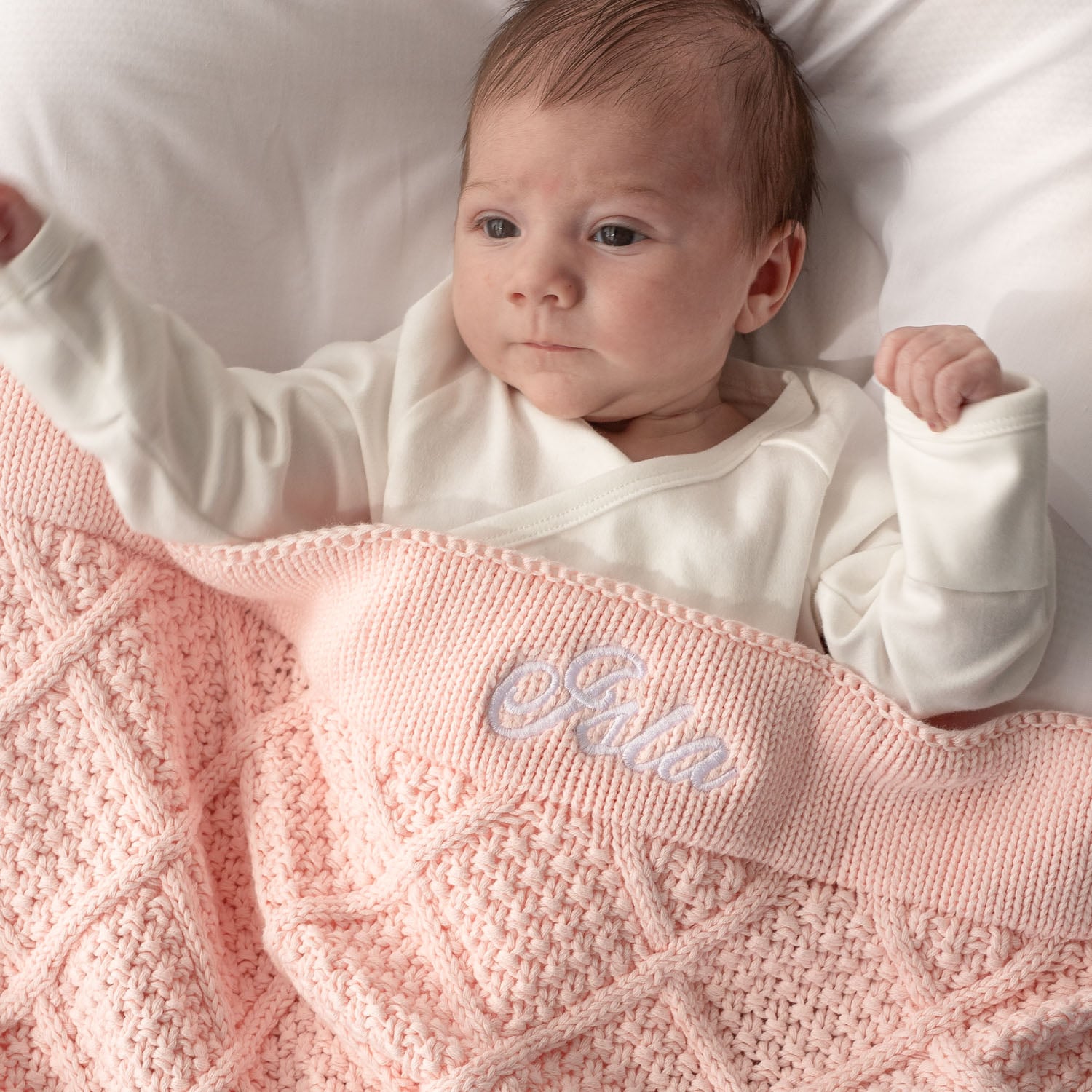 Baby lying on white bedding, wrapped in a light pink personalised knitted blanket with Isla embroidered in white.