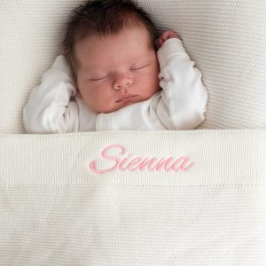 Baby laying under a white knitted cotton blanket personalised with the name Sienna with pink thread..