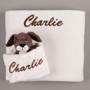 White knitted blanket embroidered with "Charlie" in brown script, laid flat with a matching brown puppy comforter embroidered "Charlie" tucked at the corner.