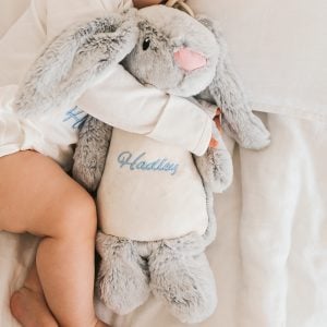 Baby cuddling a personalised grey plush bunny with blue embroidered name on its soft tummy.