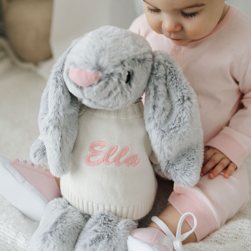 Baby in pink outfit sits beside grey plush bunny wearing a sweater embroidered with the name Ella.