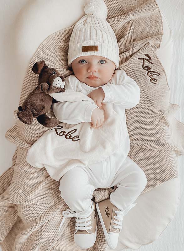 A baby with personalised beige baby blanket, puppy comforter and baby shoes.