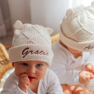 Two babies wearing cream diamond knitted beanies with ears, embroidered with the names Gracie and Archie.