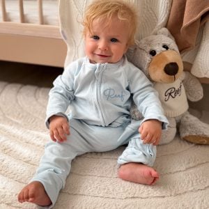 Smiling baby in personalised blue zip romper seated beside a matching embroidered teddy bear.
