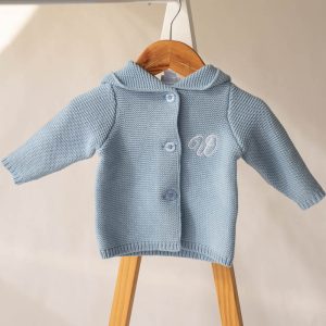 Blue knitted baby cardigan with hood and monogram, hanging on wooden hanger in soft light.