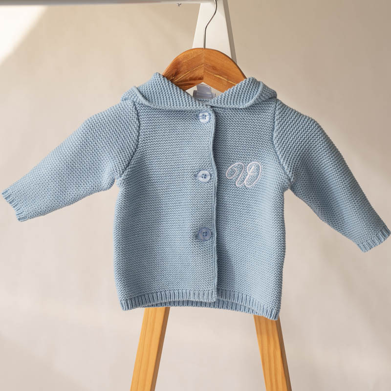 Blue Cardigan On Hanger Blue knitted baby cardigan with hood and monogram, hanging on wooden hanger in soft light.