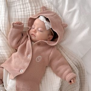 A baby sleeping in a Blush Pink Hooded Knitted Baby Cardigan embroidered with the letter O.