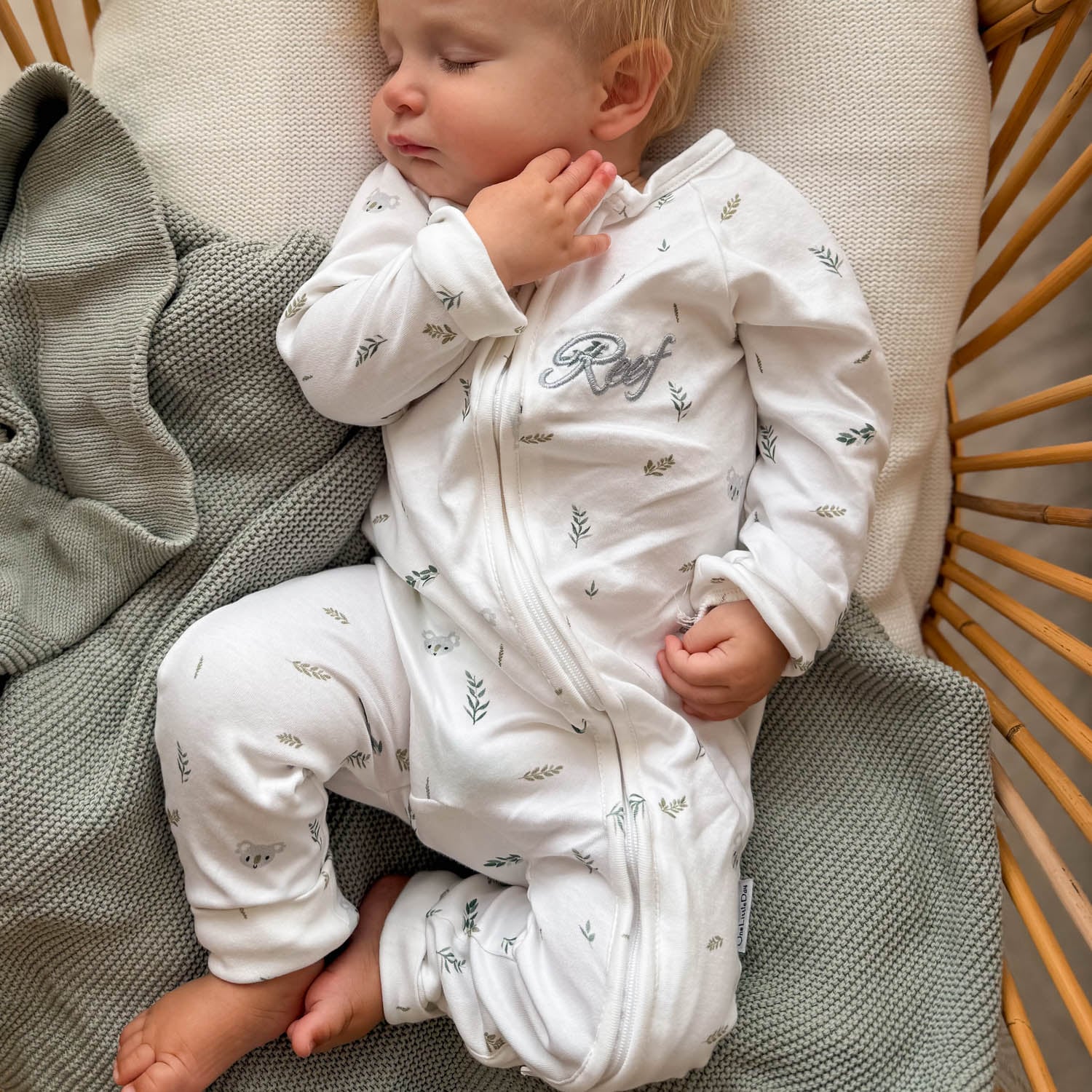 Sleeping baby in personalised white koala print zip romper in a rattan chair with sage blanket.