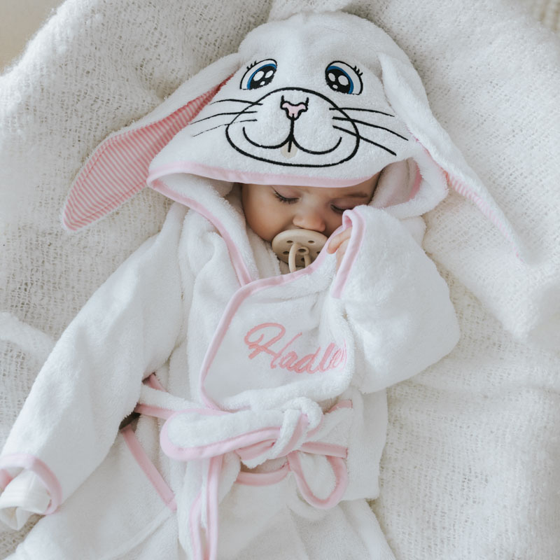 Baby sleeping in white personalised bunny robe with floppy ears and pink trim, holding a pacifier.