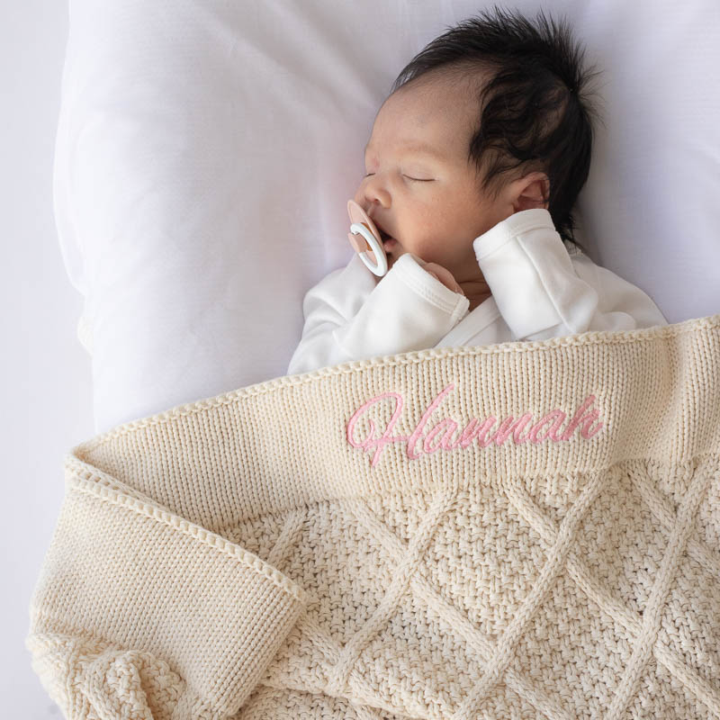 Baby Hannah laying under Cream Diamond Baby Blanket with her name embroidered in pink.