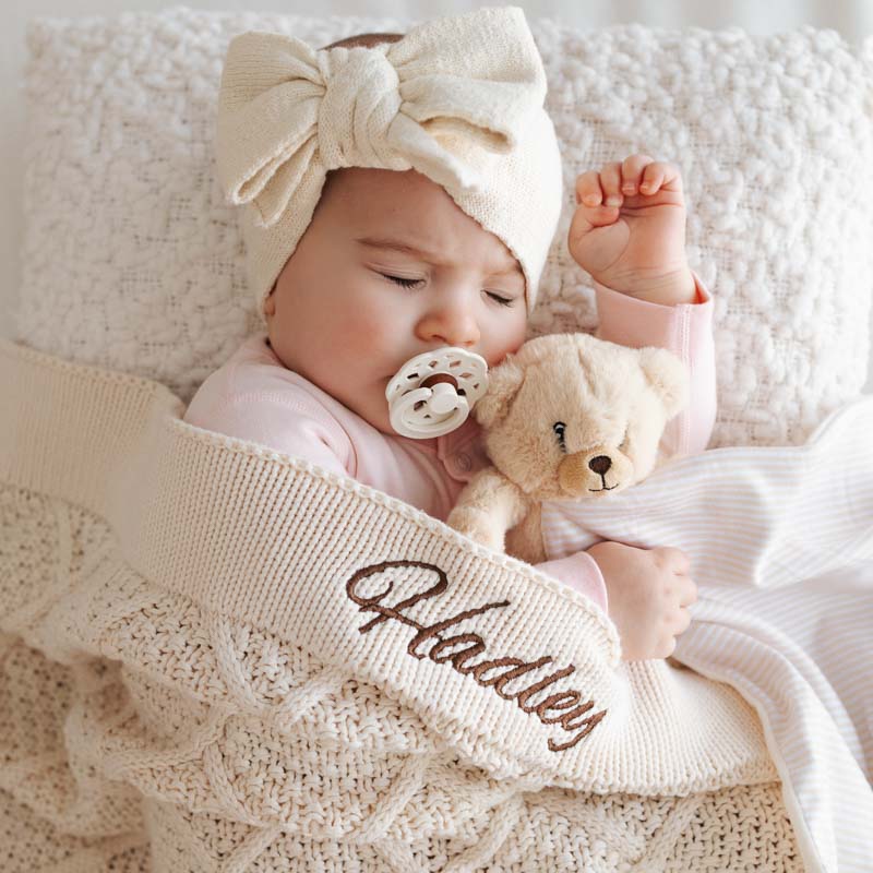 Baby sleeping with a, cream bow headband, teddy bear, and personalised cream diamond-knitted blanket embroidered with the name “Hadley.