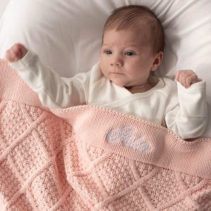 Baby lying on white bedding, wrapped in a light pink personalised knitted blanket with Isla embroidered in white.