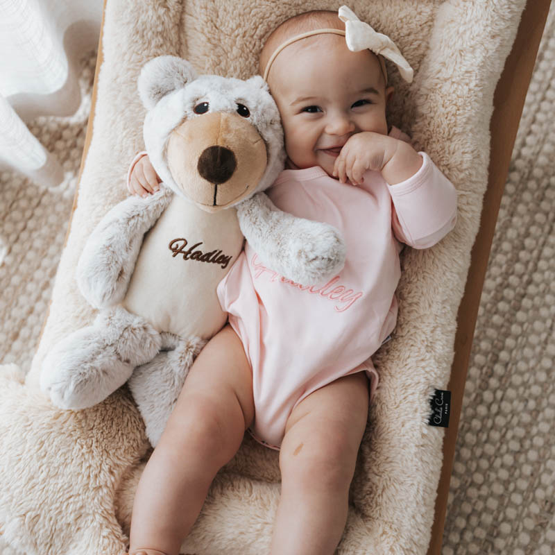 Smiling baby cuddling a personalised teddy bear while wearing a pink organic cotton personalised romper.