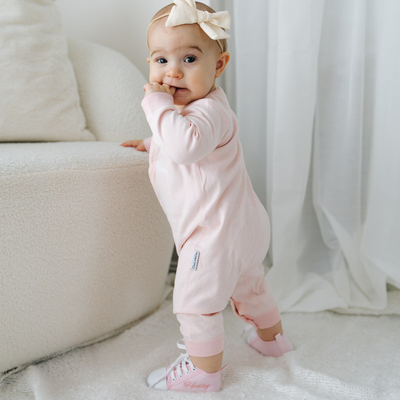 Baby girl wearing a personalised pink onesie and embroidered pink shoes, standing with a bow headband.