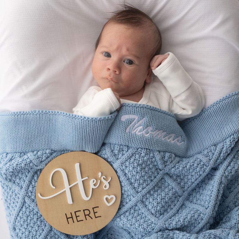 Newborn baby wrapped in blue knitted blanket with “He’s Here” birth announcement disc.
