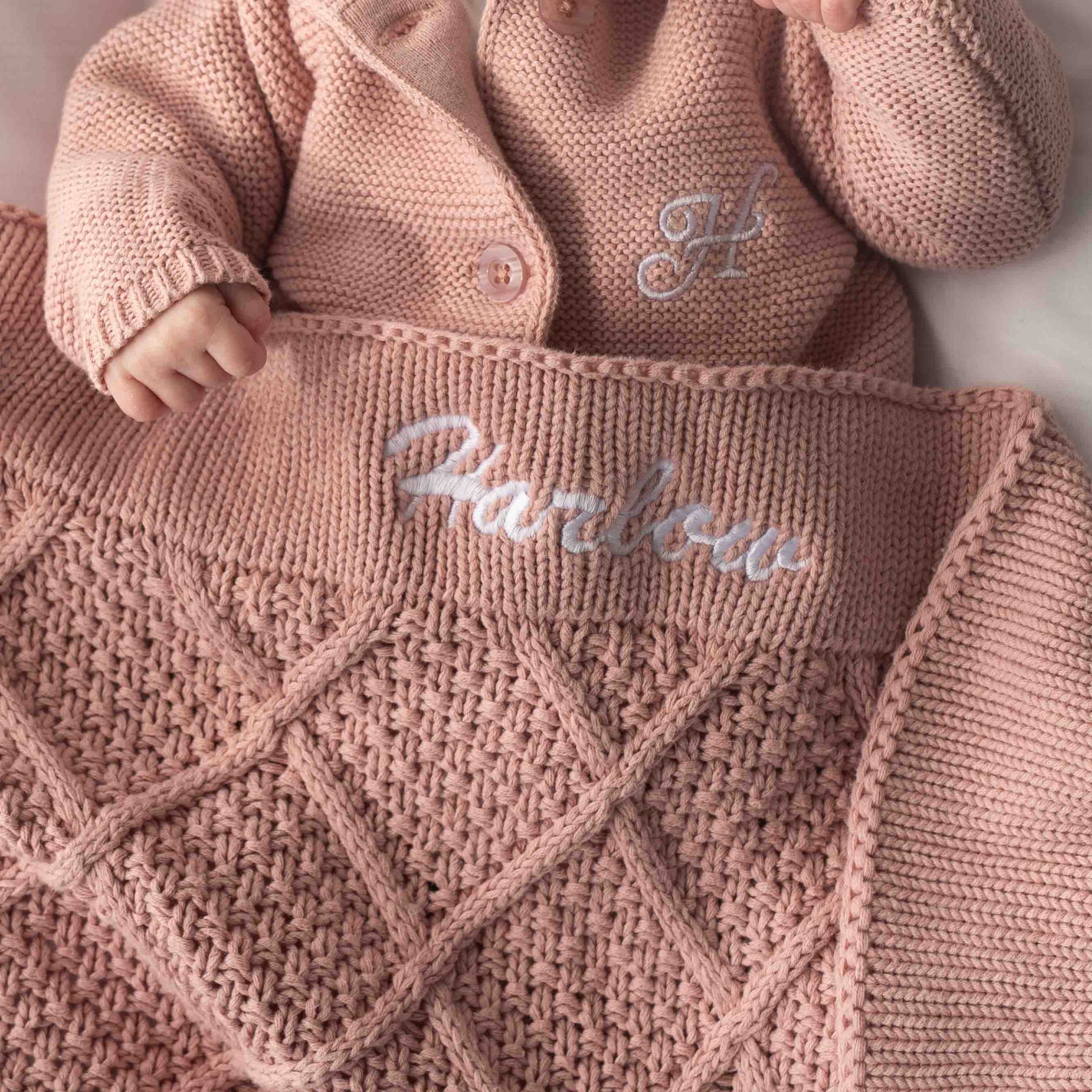 Baby wearing blush pink knitted cardigan with matching personalised diamond-pattern blanket embroidered with name Harlow.