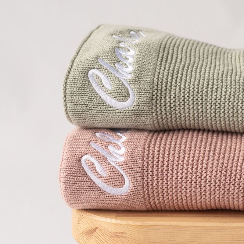 Green sage and blush pink personalised knitted baby blankets with embroidered names, stacked neatly on a wooden surface.