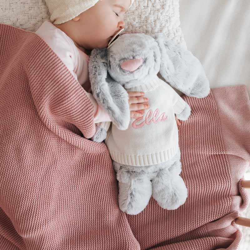 Baby sleeping under blush pink blanket, cuddling grey bunny wearing personalised jumper with “Ella” embroidered in pink.