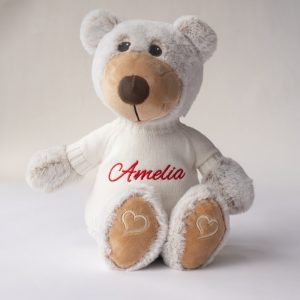 A plush teddy bear wearing a white sweater embroidered with “Amelia” in red and hearts on its feet.