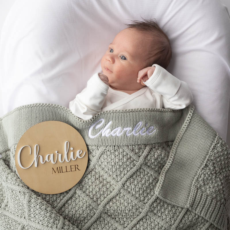 Newborn wrapped in personalised sage green knitted blanket with matching 3D birth announcement disc.