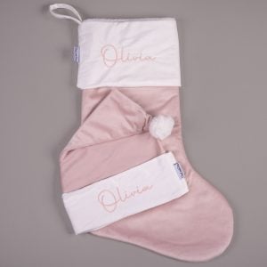 Light pink velvet personalised baby Christmas stocking and matching Santa hat with white cuffs, pompom and pink embroidered script name on grey background.