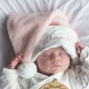 Sleeping baby wearing a fluffy pink and white Christmas hat embroidered with the name “Freya.”