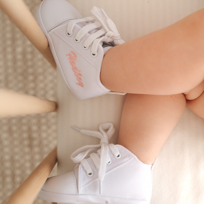 Baby wearing personalised white shoes with embroidered name, lying in a cot with crossed legs.