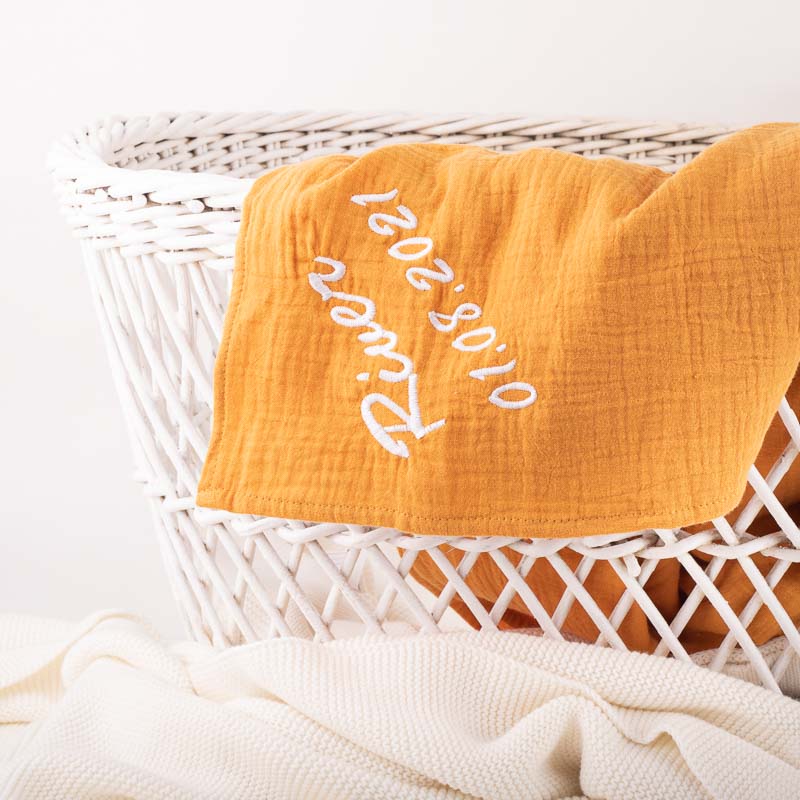 Mustard muslin baby wrap with embroidered name draped over white wicker basket.