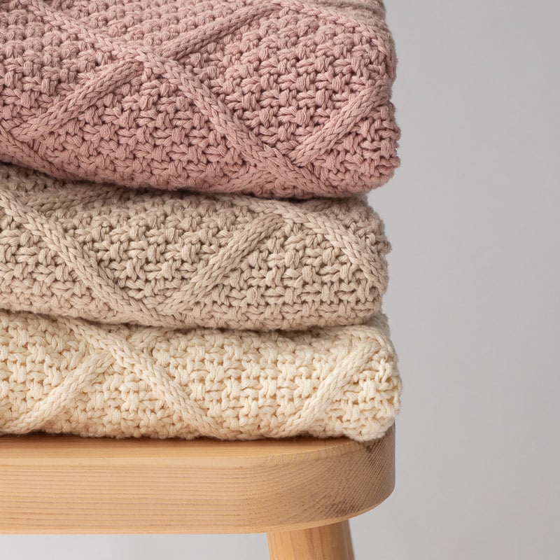 Softly knitted diamond baby blankets in blush, beige, and cream stacked on a wooden stool.