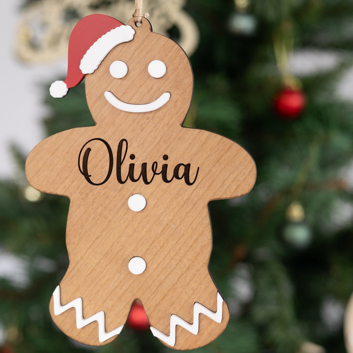 Personalised wooden gingerbread man Christmas ornament with Olivia engraved and red Santa hat hanging on tree.